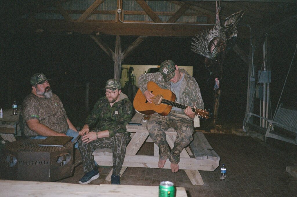 A man plays a guitar while sitting next to two hunters in camo.