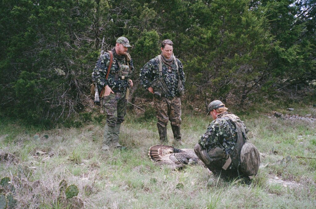 Three hunters in camo stand around a dead turkey.