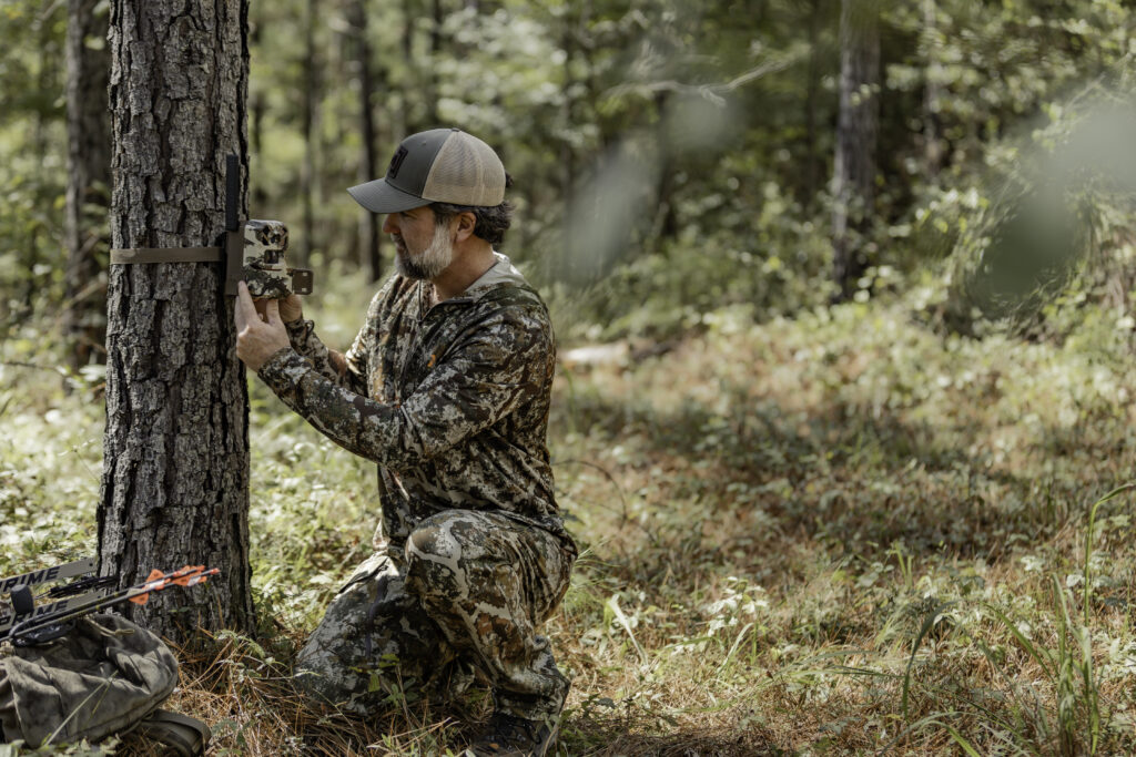 A man places a trail camera on a tree in the woods.