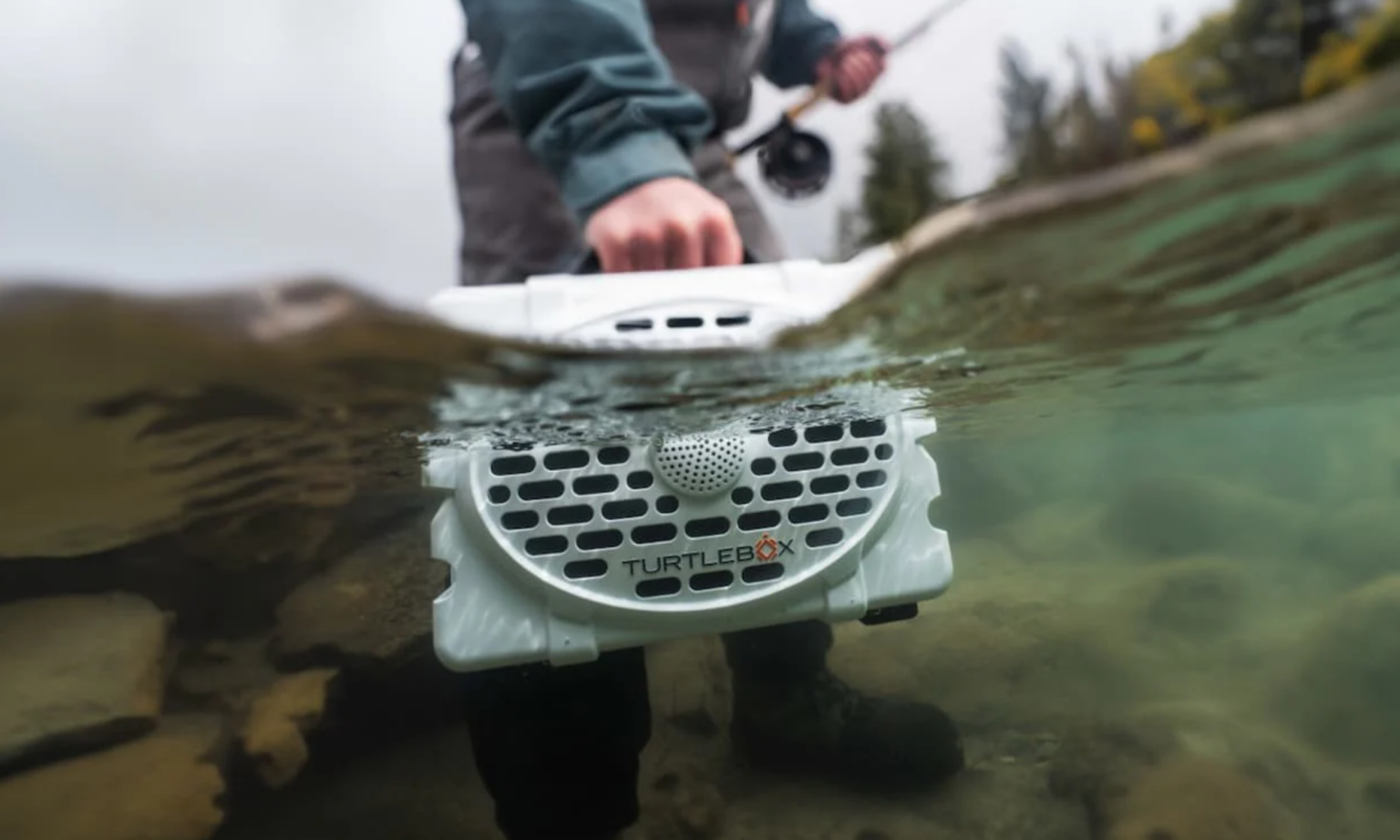 Turtlebox waterproof outdoor speaker half-submerged in river while man fly fishes nearby