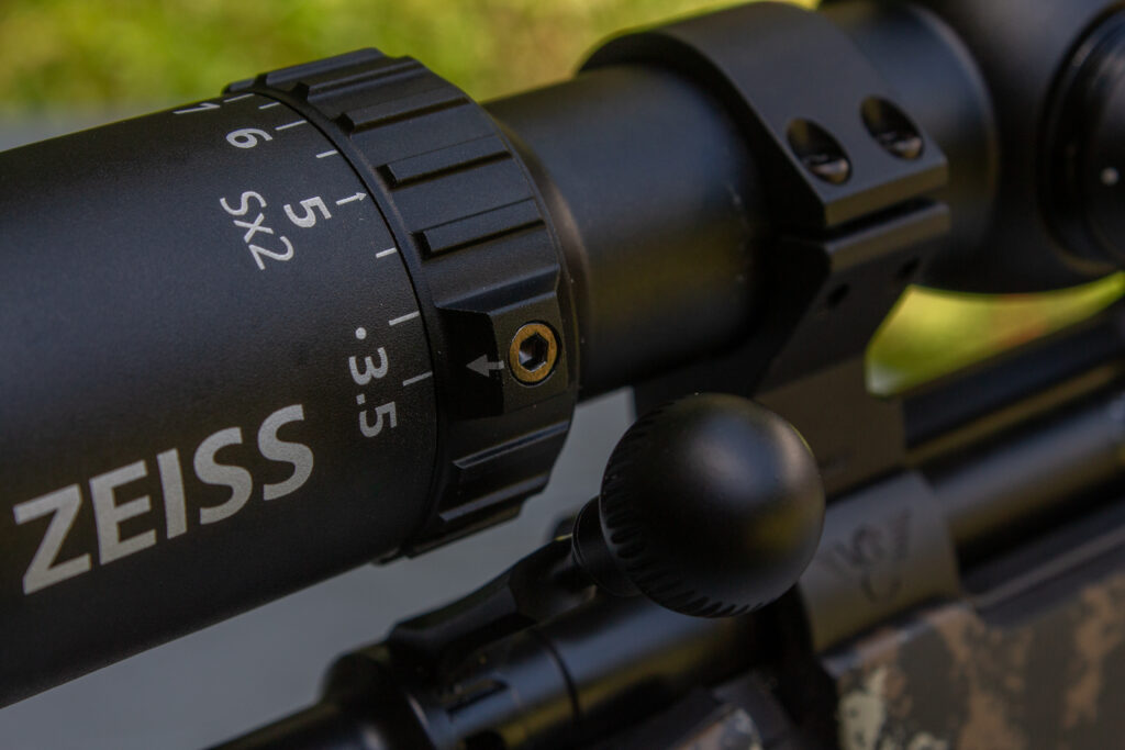 Detail of a riflescope mounted on a rifle showing the magnification adjustment ring and power indicator in relation to the rifle’s open bolt.