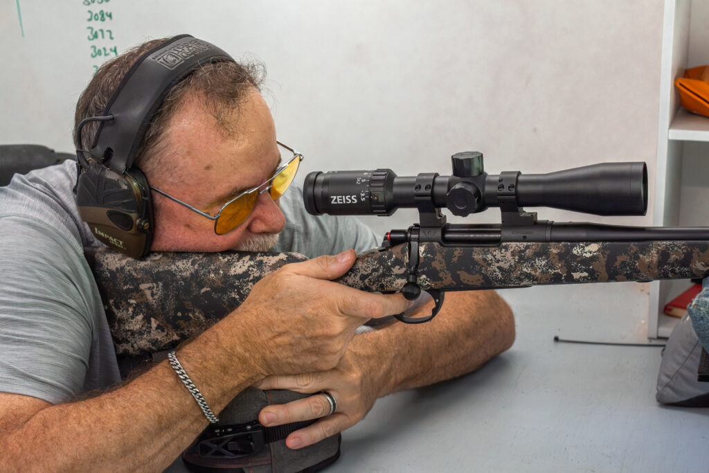 Shooter, shooting a rifle outfitted with a riflescope from a benchrest with sandbag support from inside a shooting house.