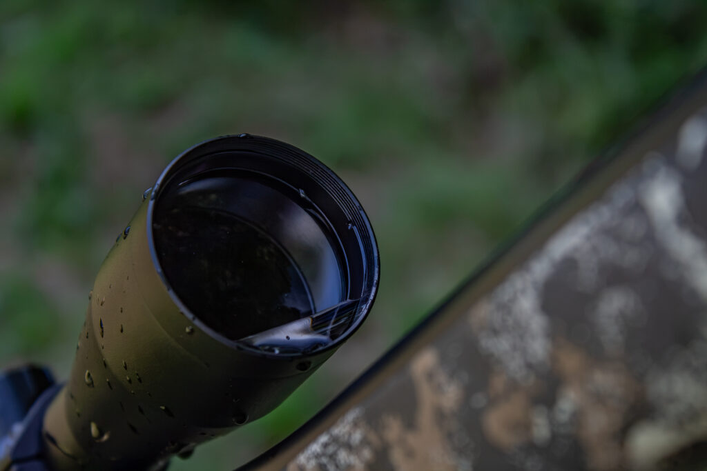 The objective lens of a riflescope positioned at an angle with water pooled at the bottom but with no water beads on the lens.