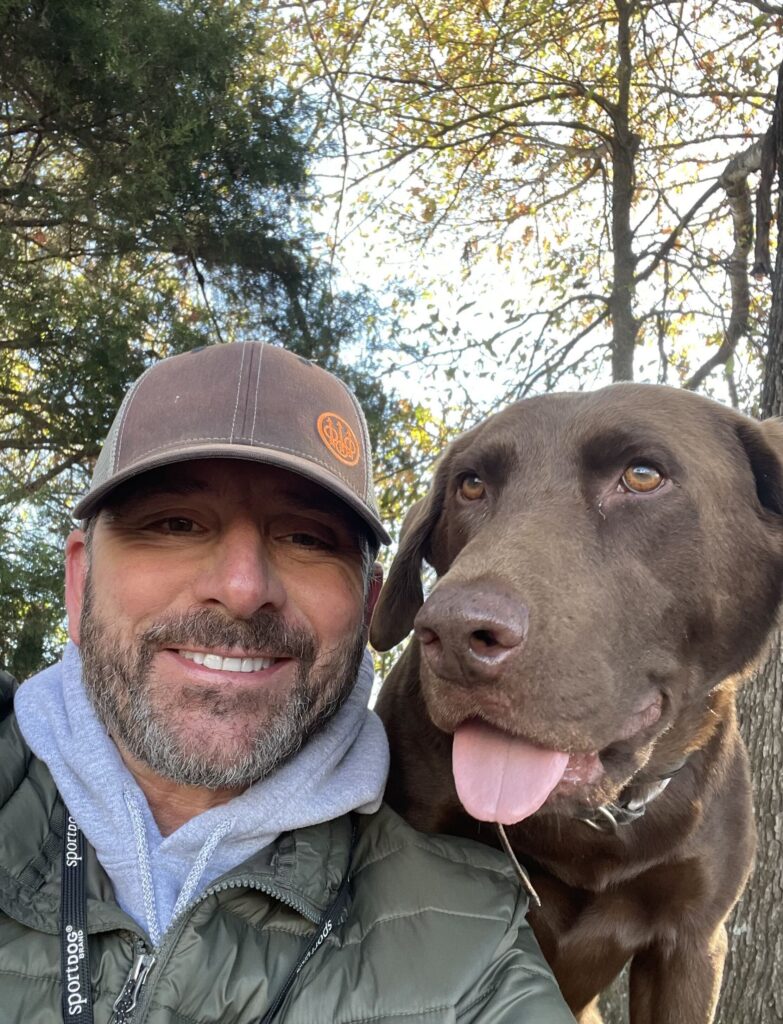 A man in a hat and green shirt smiles next to a Chocolate lab