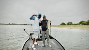 Two fishermen stand on a fishing boat. One is holding a large bass