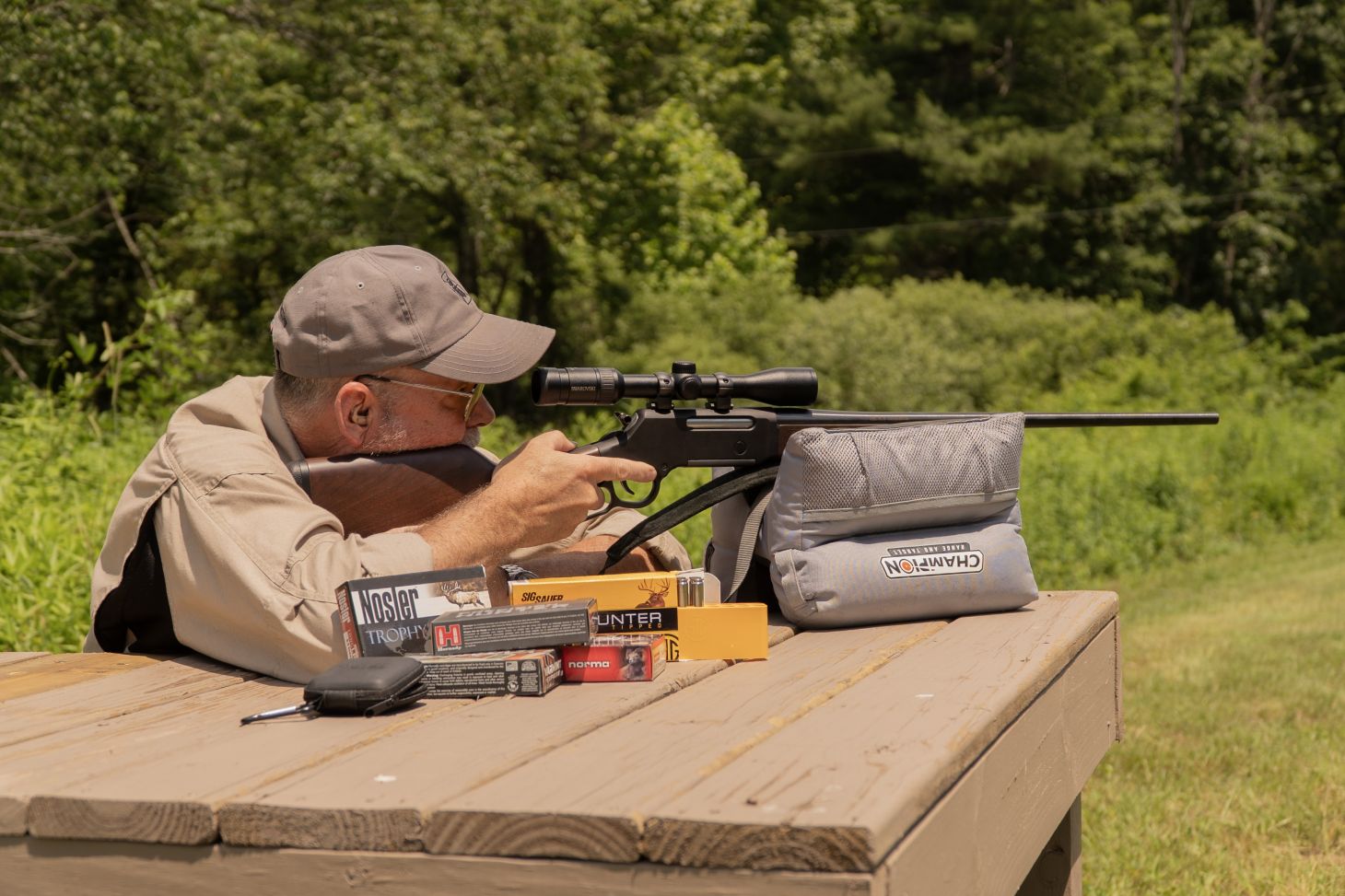 A shooter fires a scoped rifle off sand bags from a bench rest, with boxes of ammo nearby. 