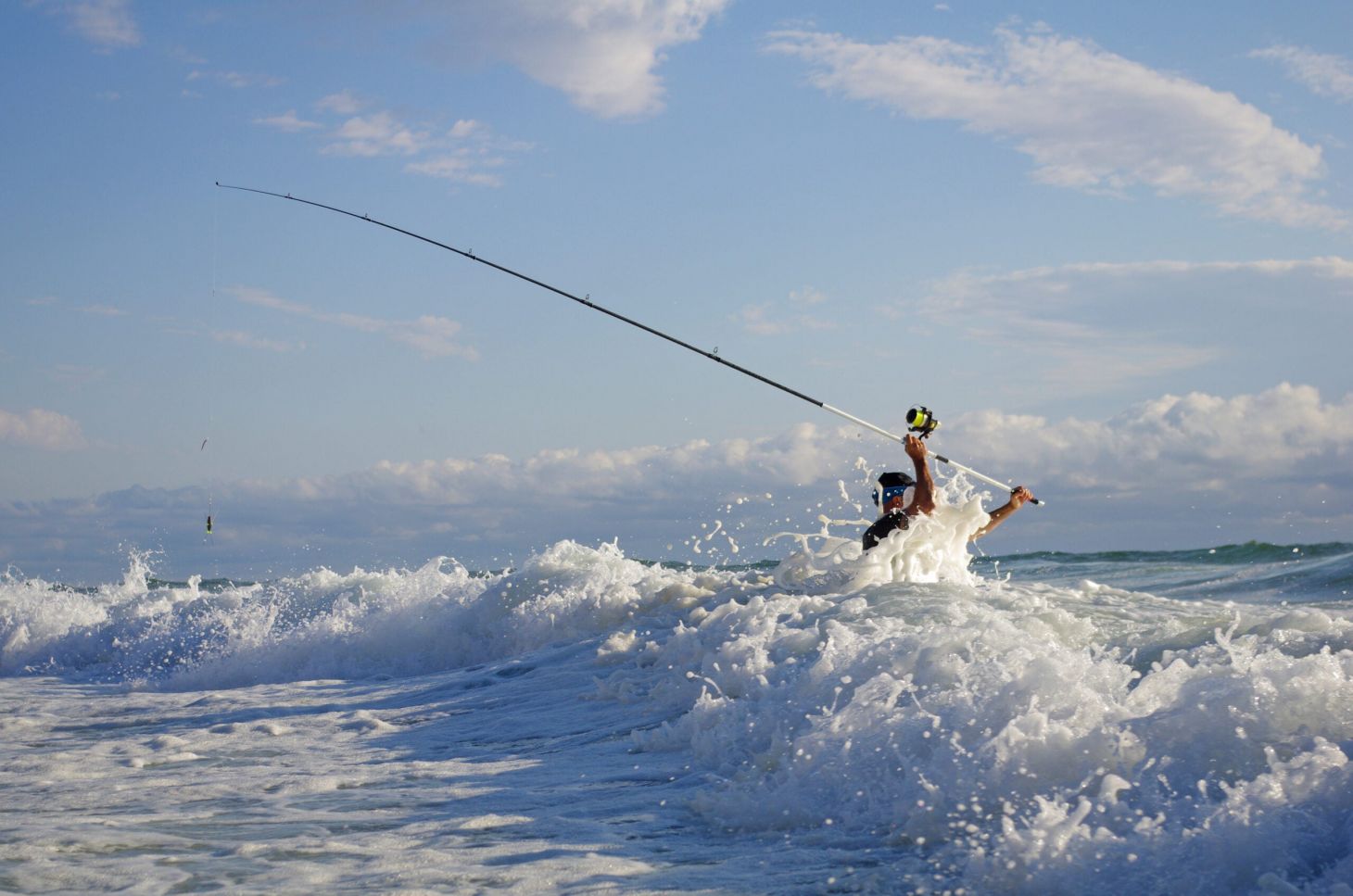A fisherman stands in front of breaking wave with a fishing rod in the air.