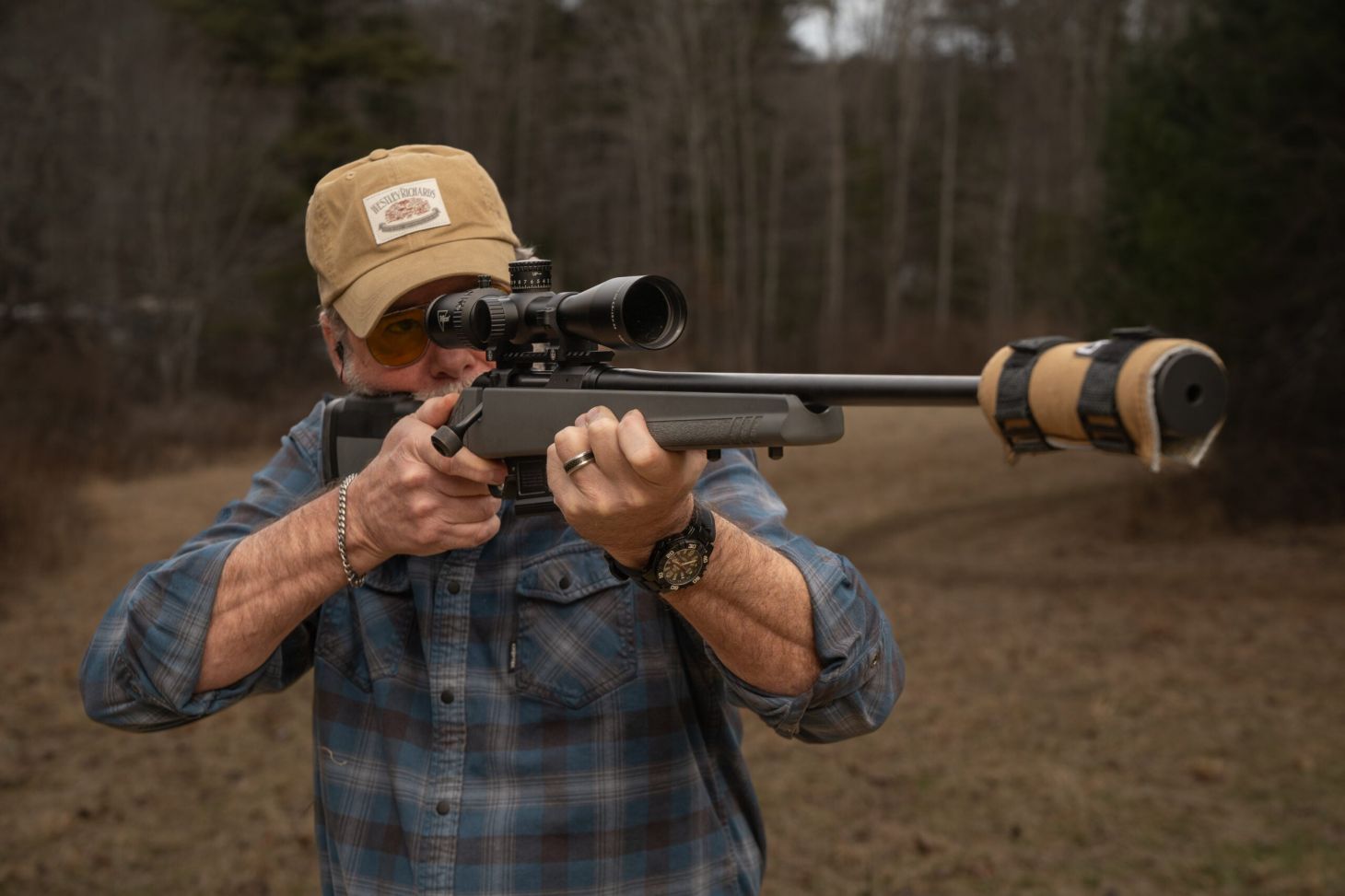 Shooter fires a rifle with silencer, field and woods in background