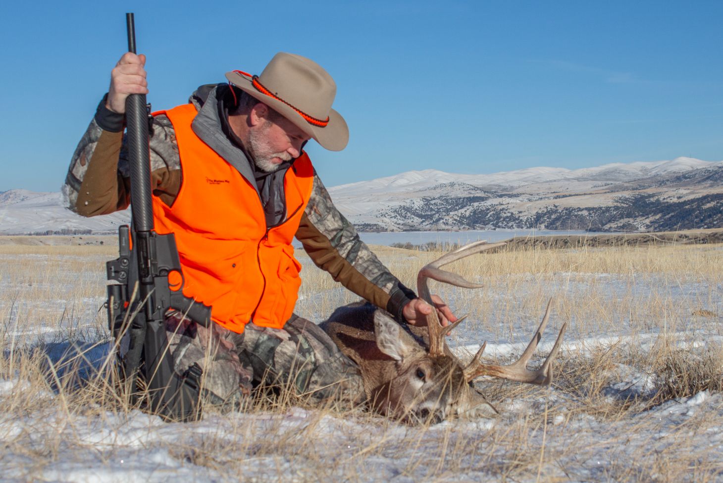 A hunter kneels next to a whitetail buck in a field. 