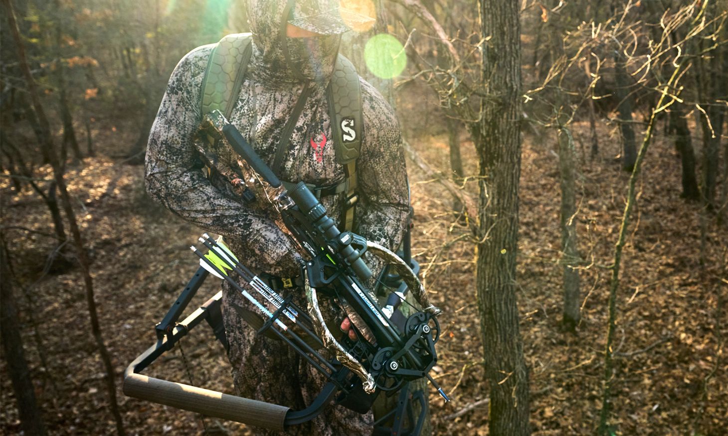 A hunter in a tree stand in the woods holding a Excalibur Hybrid-X crossbow.