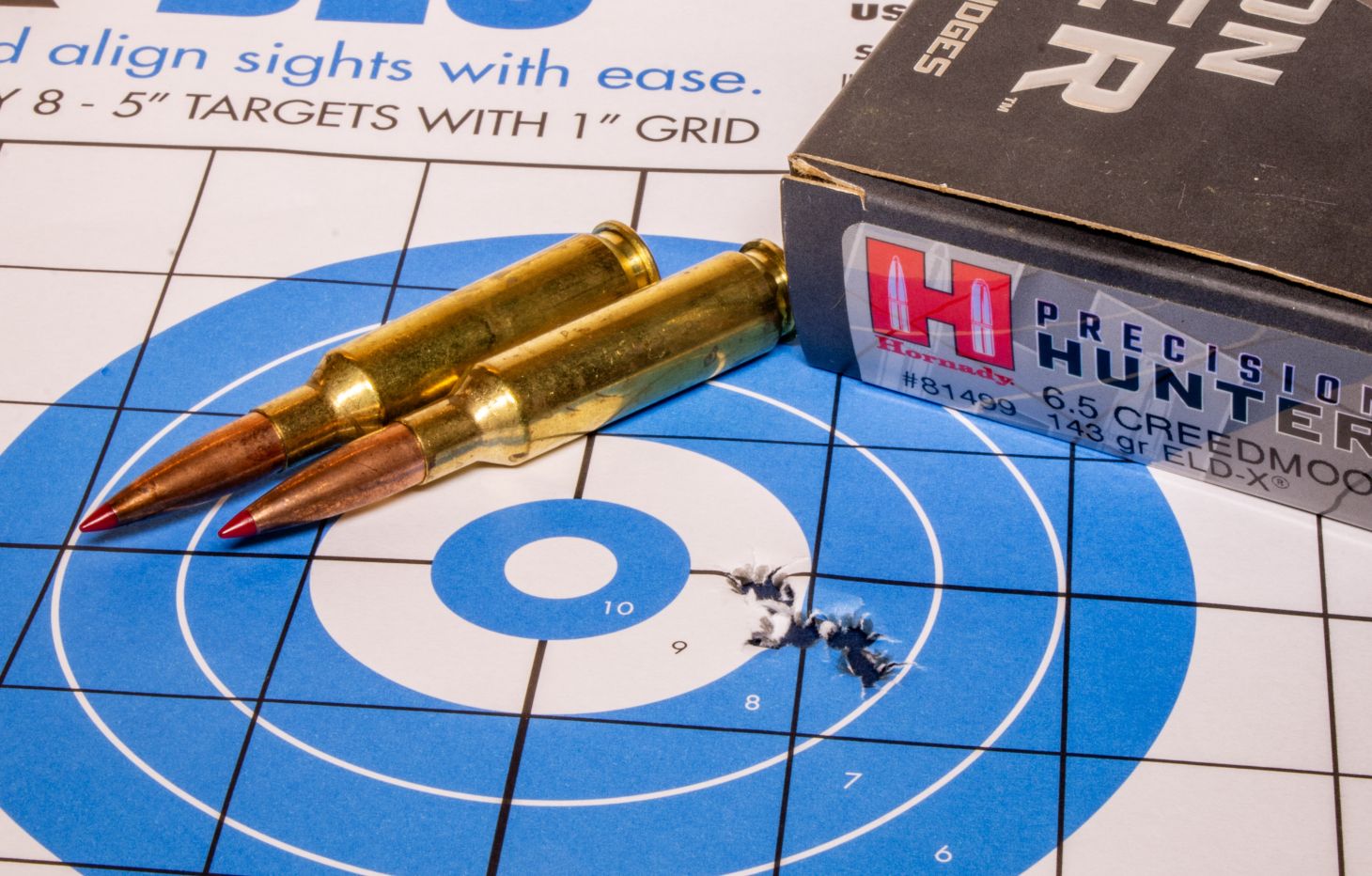 A rifle target with five bullet holes in it, with a box of ammo and two loose cartridges. 