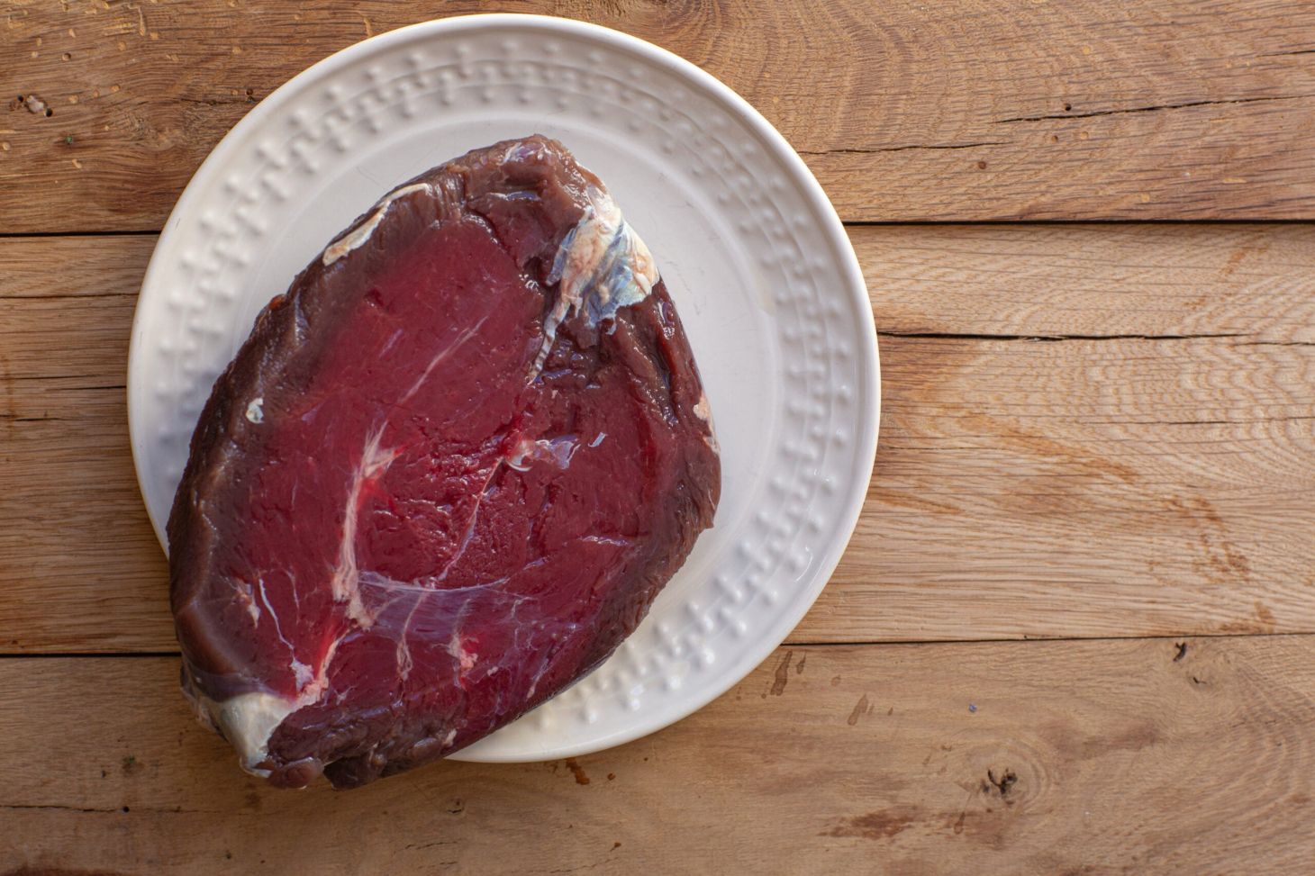 A slice of raw venison sits on a white plate resting on a wooden table.