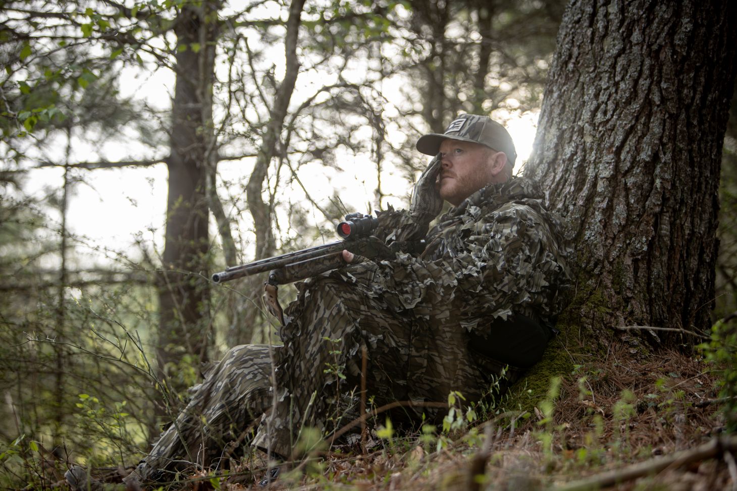 A hunter with a turkey call sits against a tree and uses a turkey mouth call. 