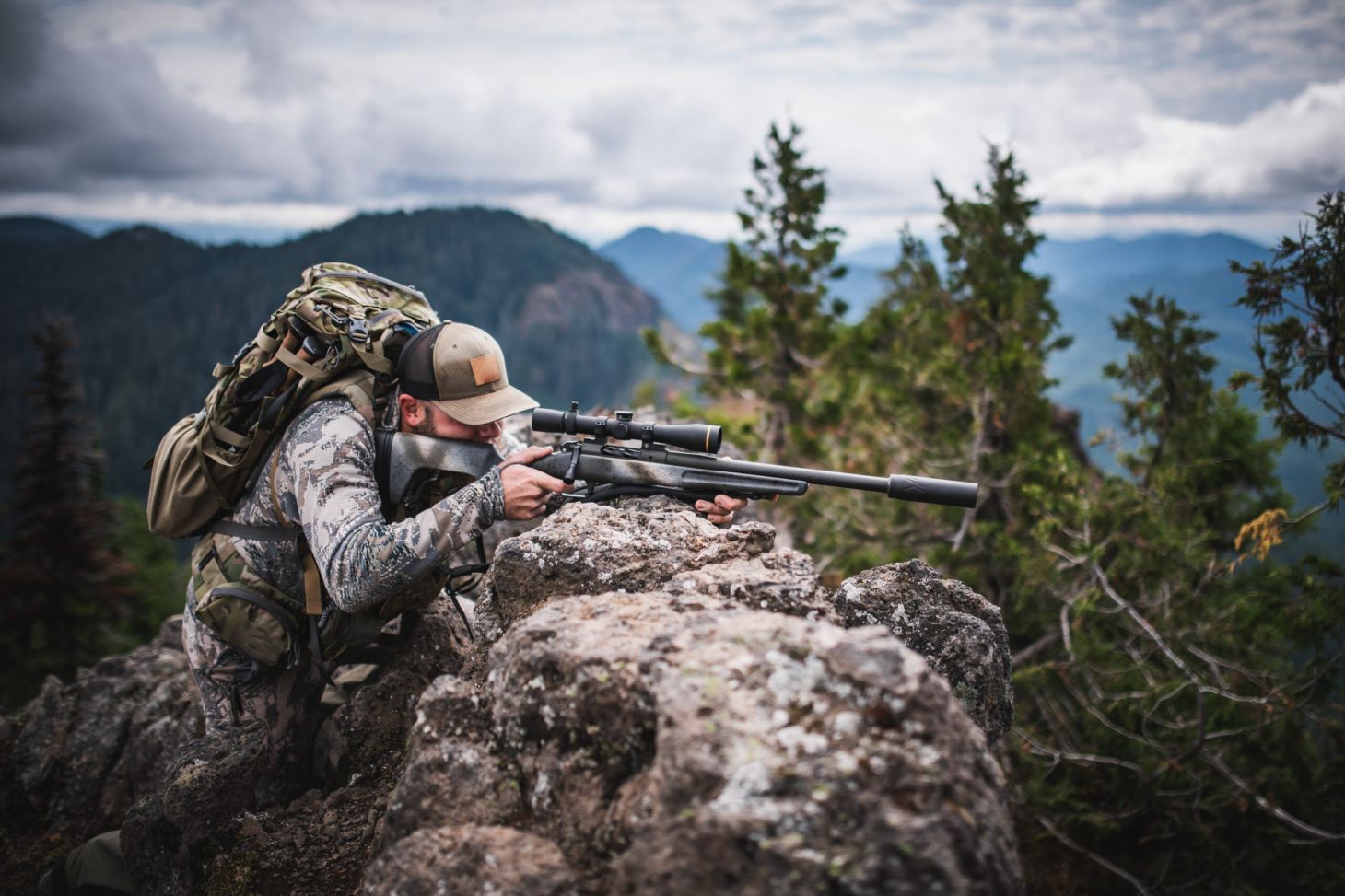 A hunter shoots a suppressed rifle from a rock outcropping on a mountain