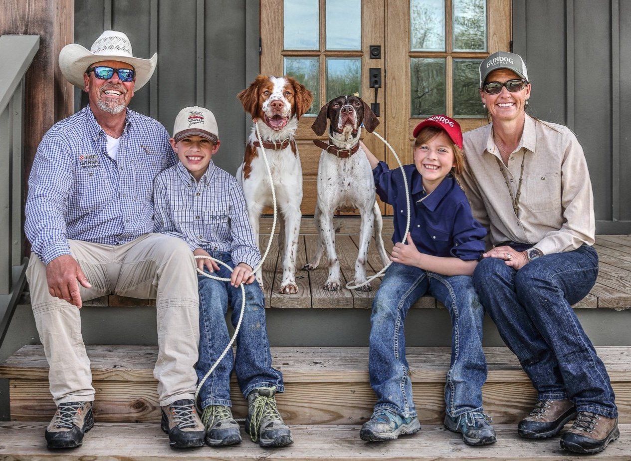 A man, woman, and two kids sit on a portch with a pair of bird hunting dogs.