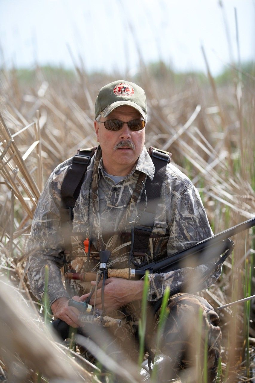 Duck hunter sits in tall reeds while holding a shotgun